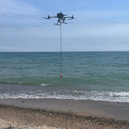 Drone flying low in sky at the coastline of a large body of water. A cord hangs from the drone into the water.