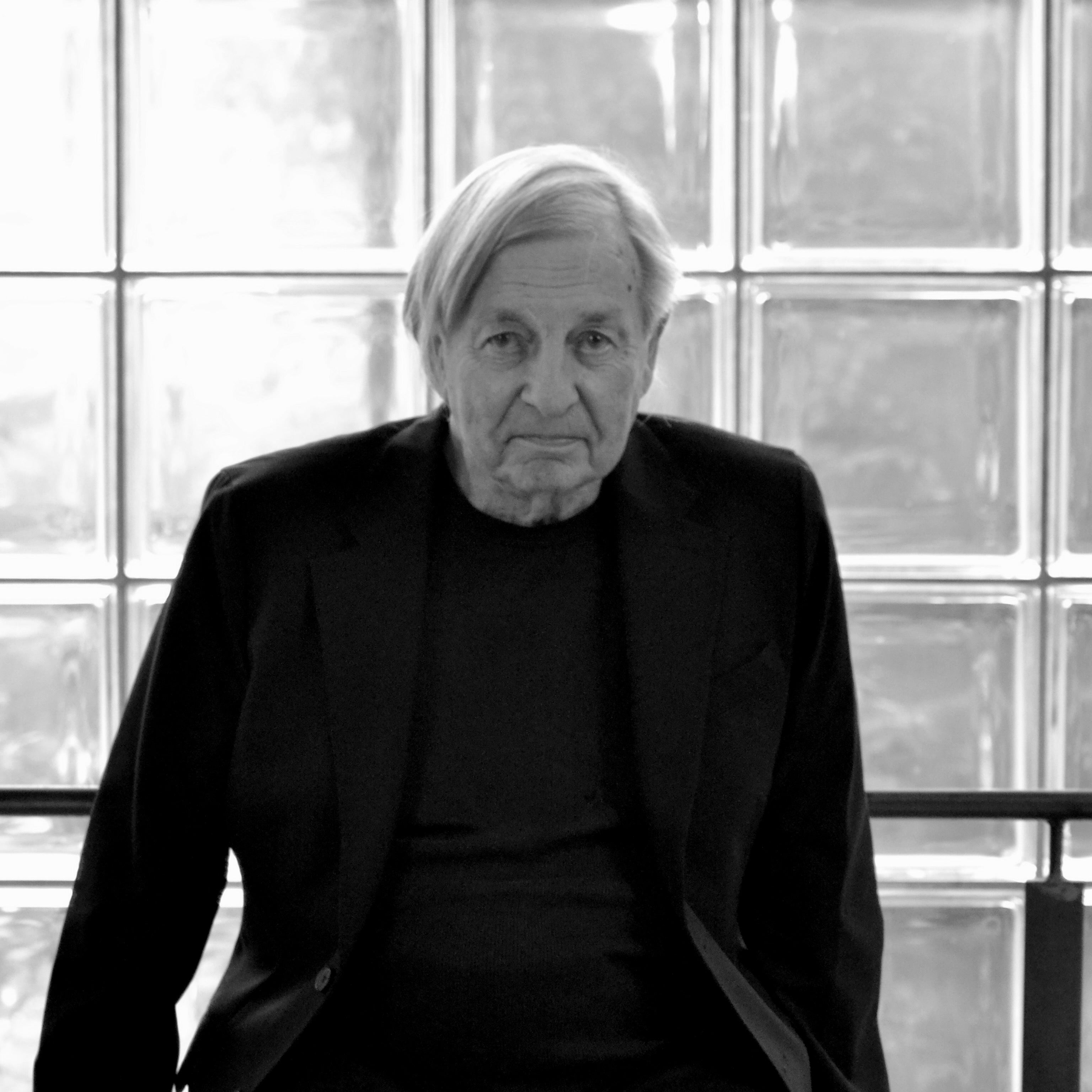 Black and white portrait of a man dressed in black standing in front of a wall of glass blocks.