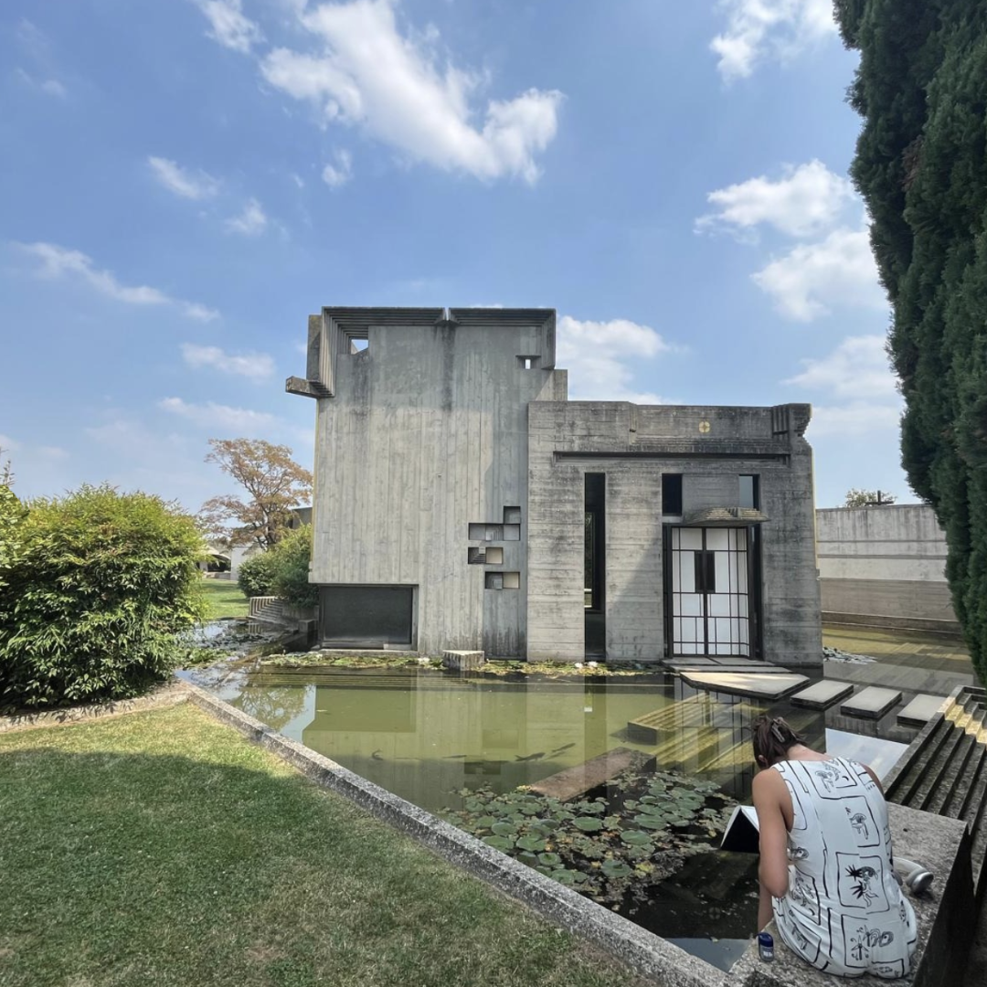 woman sketching, seated in front of a pond in front of building