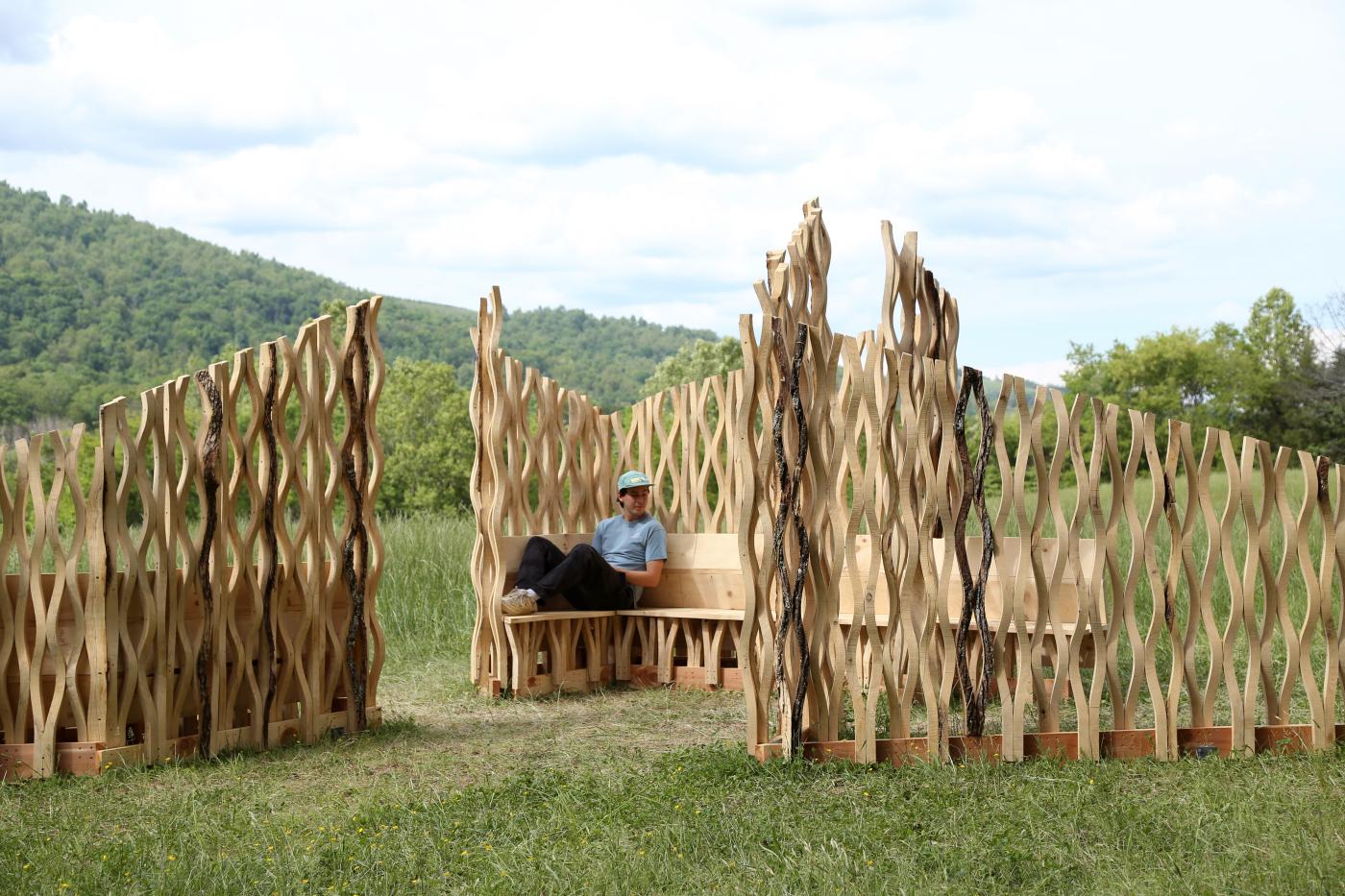 Person sitting in a bespoke wooden enclosure with curvy timber members in a hilly landscape