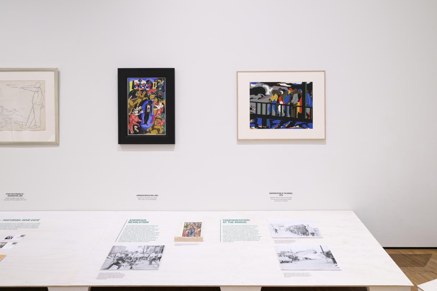 paintings on a white wall above a white floor shelf with texts and photos
