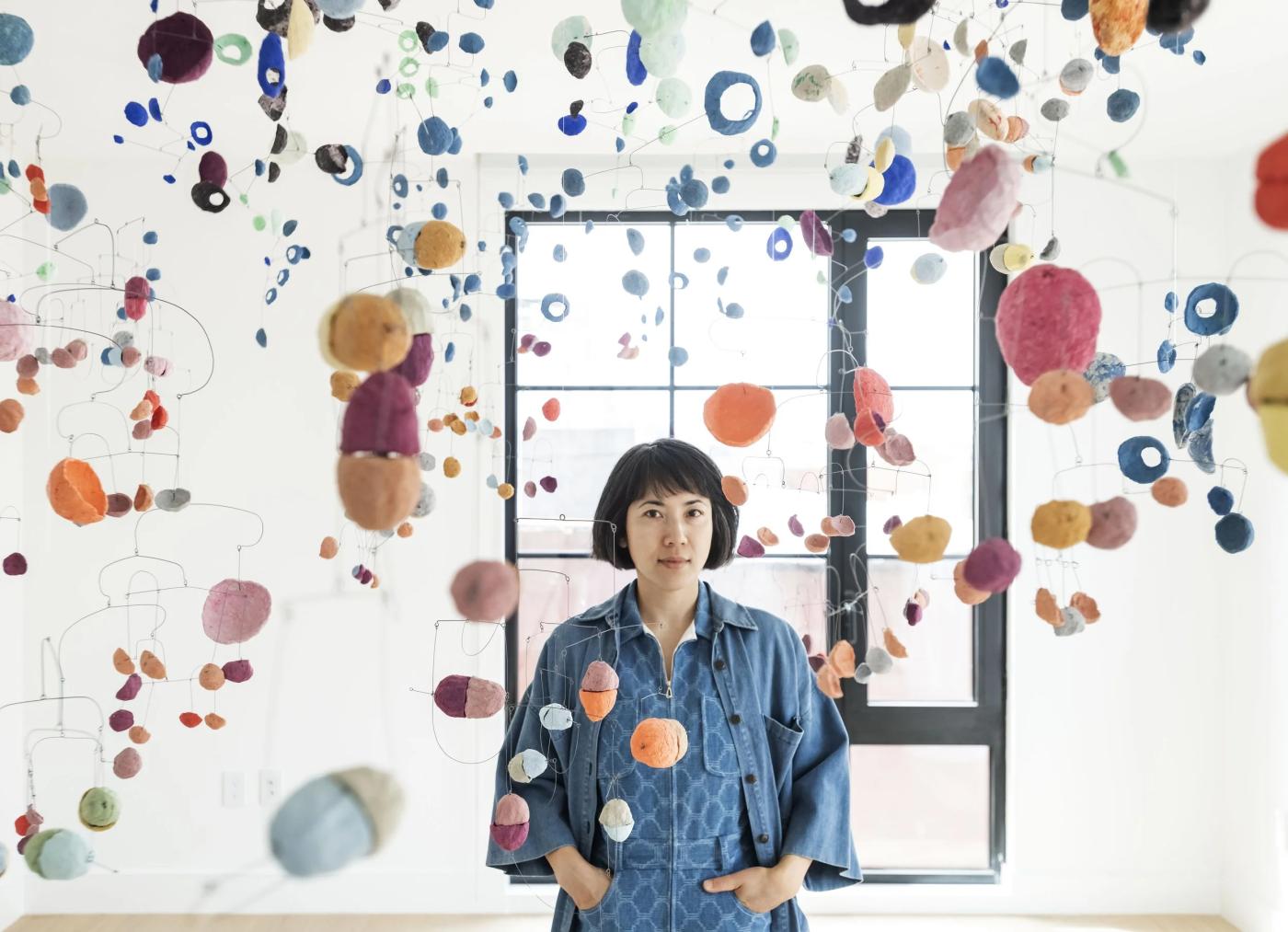 Portrait of woman standing in white room with colorful mobiles in abstract shapes hung around her